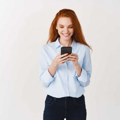 Young redhead woman reading message on mobile phone and smiling. Businesswoman looking happy at smartphone screen, standing over white background.